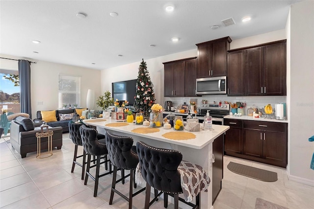 kitchen featuring a kitchen bar, appliances with stainless steel finishes, decorative backsplash, dark brown cabinetry, and an island with sink