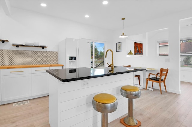 kitchen with white refrigerator with ice dispenser, a kitchen island with sink, a kitchen breakfast bar, hanging light fixtures, and white cabinetry