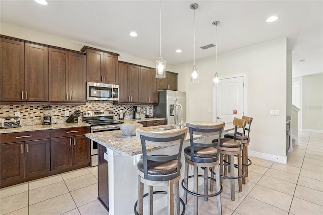 kitchen featuring pendant lighting, a center island with sink, appliances with stainless steel finishes, light stone counters, and dark brown cabinetry