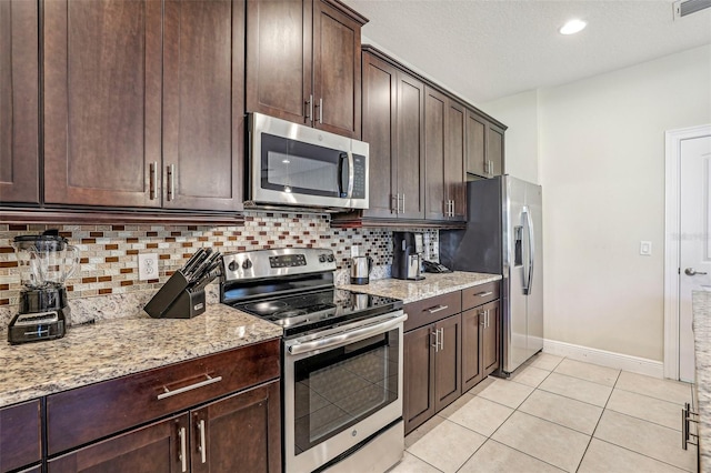 kitchen featuring dark brown cabinetry, light stone countertops, stainless steel appliances, decorative backsplash, and light tile patterned floors