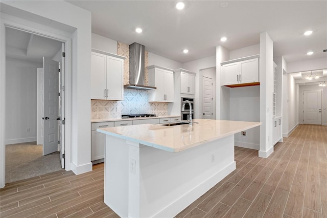 kitchen with an island with sink, white cabinets, backsplash, and wall chimney range hood