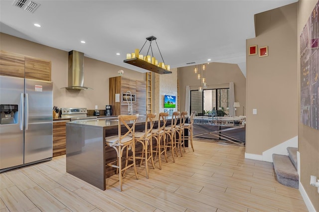 kitchen featuring hanging light fixtures, light hardwood / wood-style flooring, wall chimney exhaust hood, appliances with stainless steel finishes, and a kitchen bar