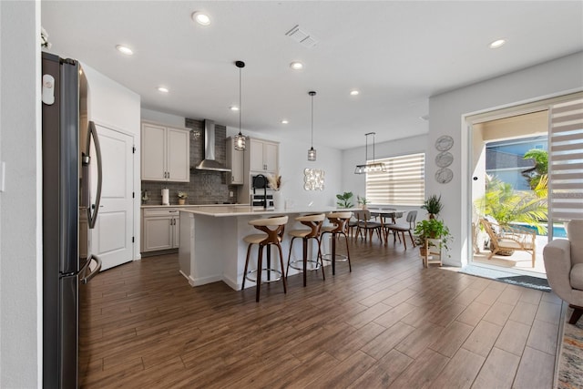 kitchen with pendant lighting, a center island with sink, wall chimney range hood, dark hardwood / wood-style floors, and stainless steel refrigerator