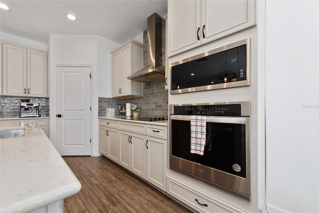 kitchen featuring dark wood-type flooring, wall chimney range hood, tasteful backsplash, light stone counters, and appliances with stainless steel finishes