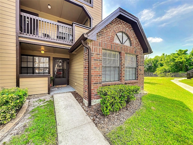 view of exterior entry featuring brick siding, a lawn, a balcony, and fence