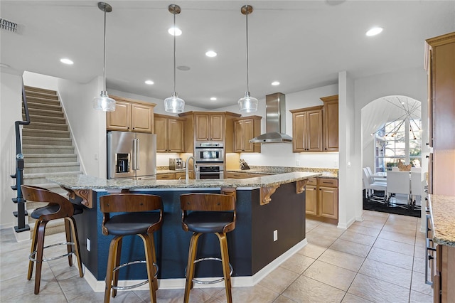 kitchen featuring decorative light fixtures, high quality fridge, a large island with sink, light stone countertops, and wall chimney range hood