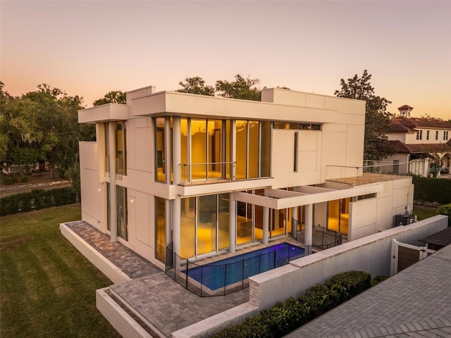 back house at dusk with a balcony, a patio area, and a lawn