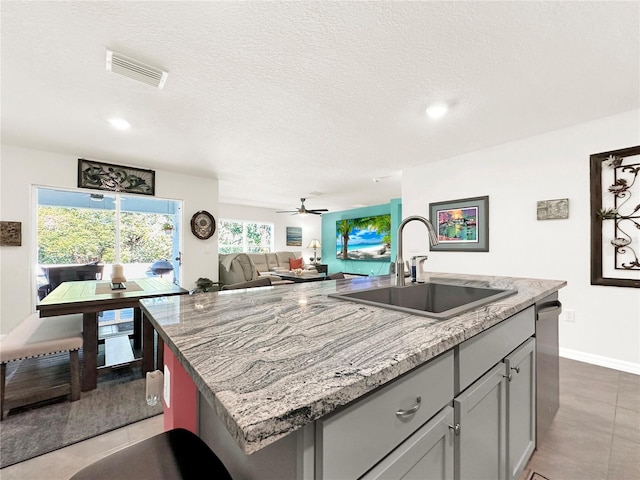 kitchen with gray cabinetry, a kitchen island with sink, sink, ceiling fan, and light stone counters