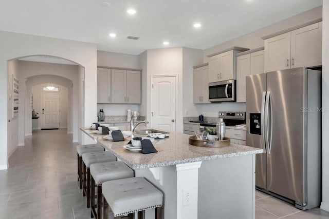 kitchen with light stone countertops, a breakfast bar, stainless steel appliances, light tile patterned floors, and a center island with sink