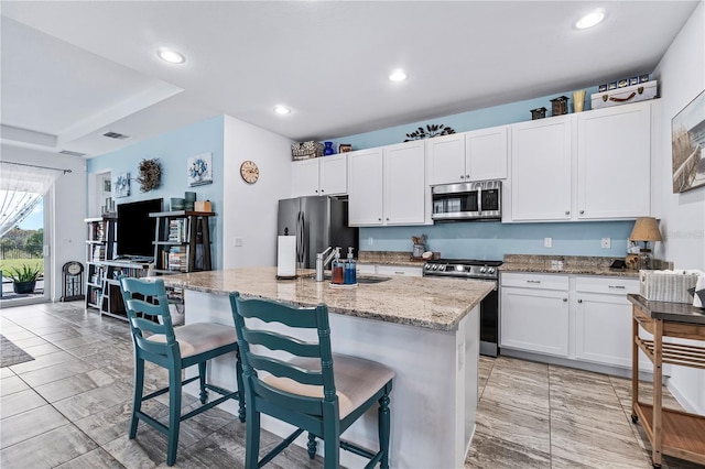 kitchen featuring a center island with sink, a kitchen breakfast bar, appliances with stainless steel finishes, light stone counters, and white cabinetry