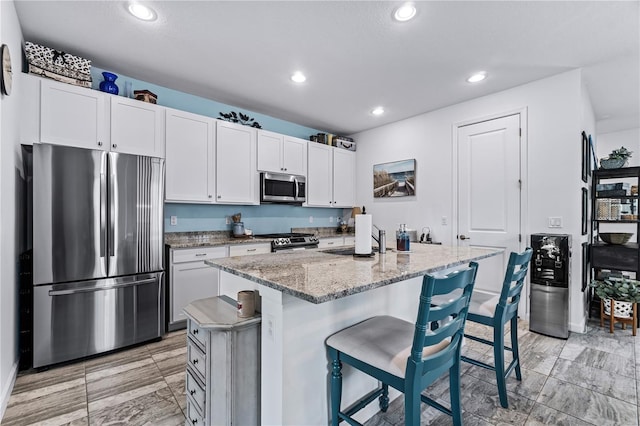 kitchen with light stone countertops, appliances with stainless steel finishes, a breakfast bar, a kitchen island with sink, and white cabinets