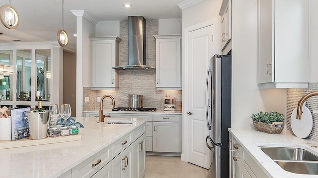 kitchen with light stone countertops, wall chimney exhaust hood, hanging light fixtures, stainless steel fridge, and ornamental molding