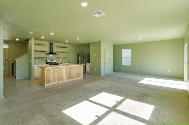 kitchen with light colored carpet, wall chimney range hood, a center island with sink, stainless steel stove, and fridge