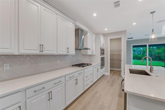 kitchen with light wood-type flooring, stainless steel appliances, sink, wall chimney range hood, and white cabinets