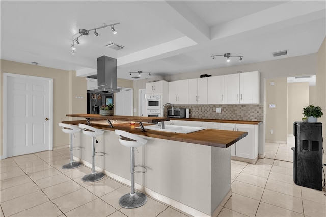 kitchen with island exhaust hood, tasteful backsplash, a center island with sink, white cabinets, and butcher block counters