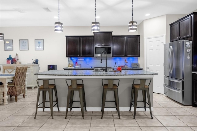 kitchen featuring dark stone counters, an island with sink, tasteful backsplash, decorative light fixtures, and stainless steel appliances