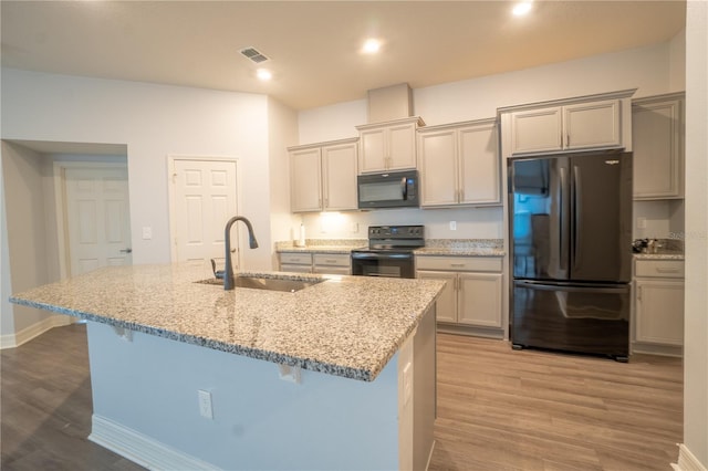 kitchen featuring a kitchen island with sink, black appliances, sink, light wood-type flooring, and light stone counters