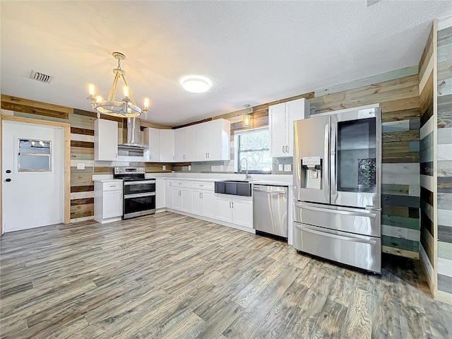 kitchen featuring white cabinetry, wall chimney exhaust hood, stainless steel appliances, pendant lighting, and wooden walls