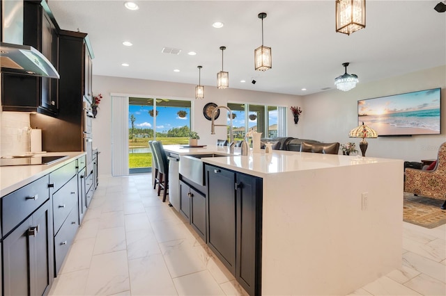 kitchen featuring a large island with sink, black electric cooktop, decorative light fixtures, and wall chimney range hood