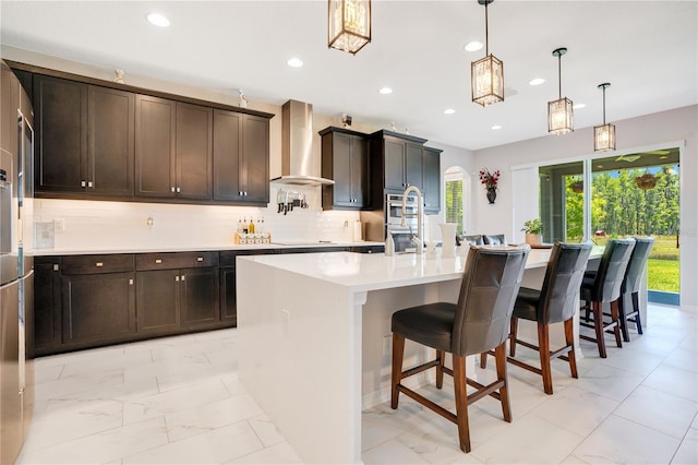 kitchen featuring wall chimney exhaust hood, hanging light fixtures, an island with sink, decorative backsplash, and black electric stovetop