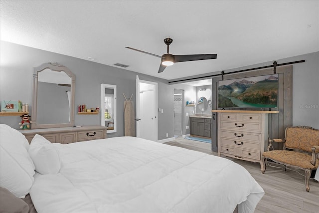 bedroom featuring a barn door, ensuite bathroom, ceiling fan, and light wood-type flooring