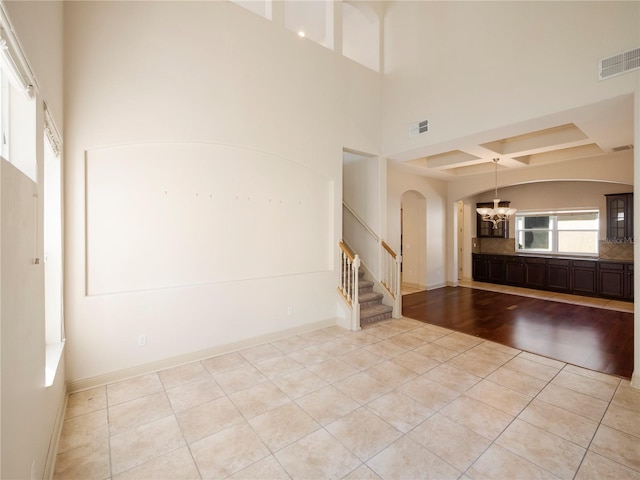 empty room with a towering ceiling, coffered ceiling, a chandelier, beamed ceiling, and light hardwood / wood-style floors