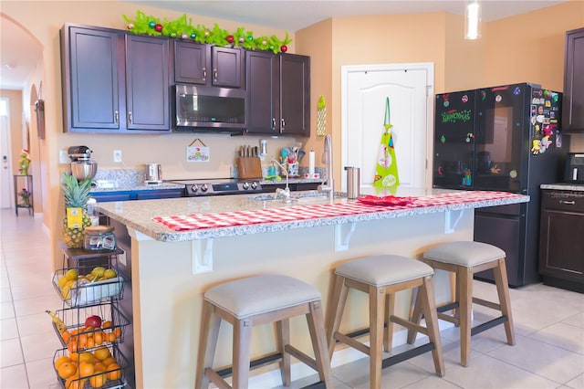 kitchen featuring black refrigerator, dark brown cabinets, light tile patterned floors, a center island with sink, and a breakfast bar area