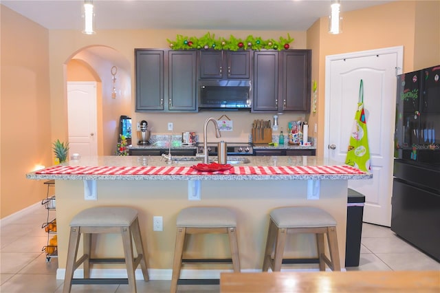 kitchen featuring black refrigerator, an island with sink, dark brown cabinets, light tile patterned flooring, and a breakfast bar area