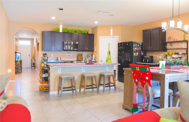 kitchen with a breakfast bar, a kitchen island with sink, black fridge, hanging light fixtures, and dark brown cabinets