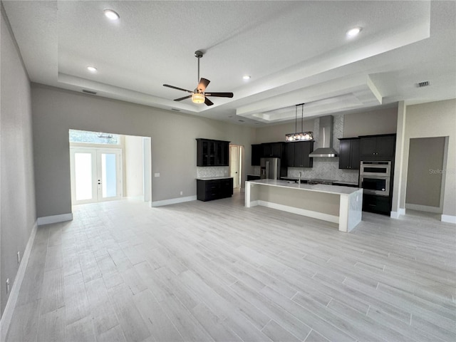 kitchen with wall chimney exhaust hood, stainless steel appliances, an island with sink, light hardwood / wood-style floors, and a tray ceiling