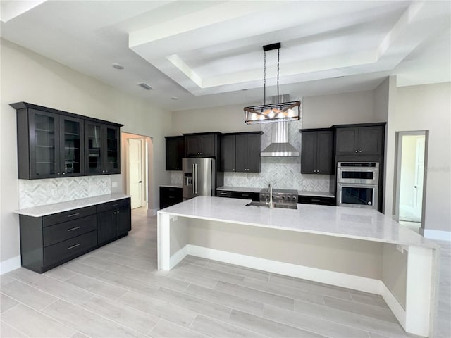 kitchen featuring a kitchen breakfast bar, a raised ceiling, wall chimney exhaust hood, tasteful backsplash, and stainless steel appliances