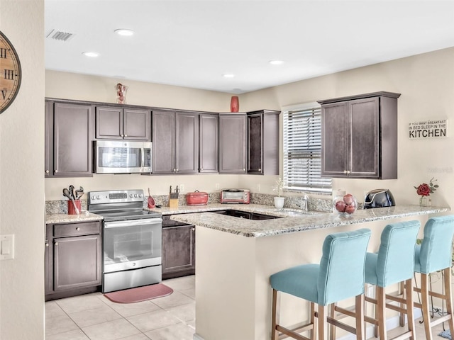 kitchen with kitchen peninsula, light stone countertops, dark brown cabinetry, stainless steel appliances, and light tile patterned floors