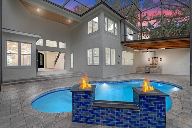 pool at dusk with a fire pit, a patio, ceiling fan, and an outdoor kitchen