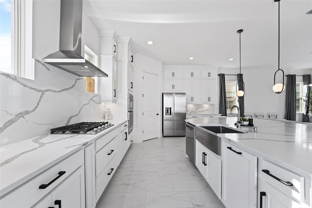 kitchen with wall chimney range hood, sink, hanging light fixtures, white cabinetry, and stainless steel appliances