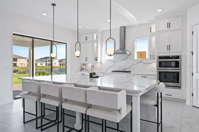 kitchen with a kitchen breakfast bar, light stone counters, wall chimney exhaust hood, decorative light fixtures, and white cabinets