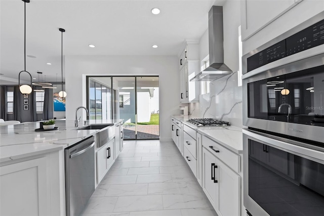 kitchen featuring white cabinets, wall chimney range hood, hanging light fixtures, appliances with stainless steel finishes, and light stone counters