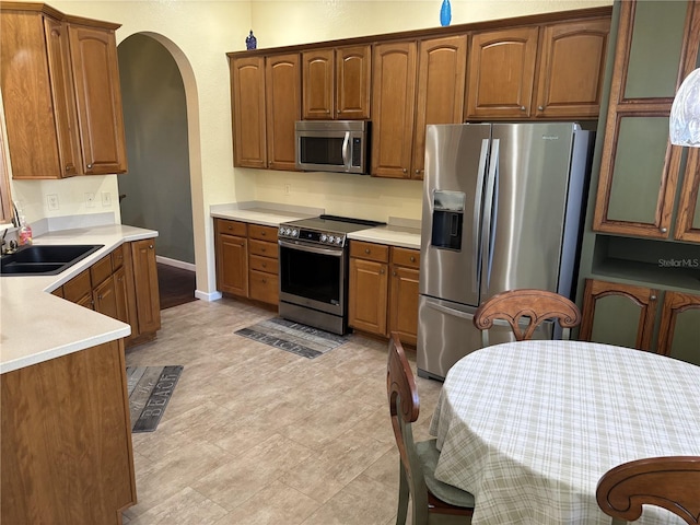 kitchen featuring arched walkways, appliances with stainless steel finishes, brown cabinets, light countertops, and a sink