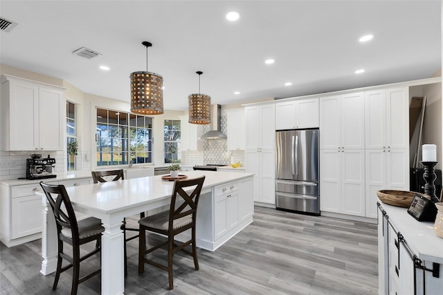 kitchen featuring white cabinets, decorative light fixtures, wall chimney range hood, and stainless steel refrigerator