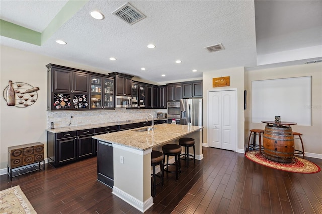 kitchen with light stone countertops, sink, dark wood-type flooring, an island with sink, and appliances with stainless steel finishes