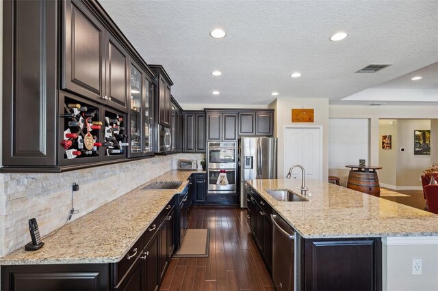 kitchen featuring sink, an island with sink, appliances with stainless steel finishes, dark hardwood / wood-style flooring, and light stone counters