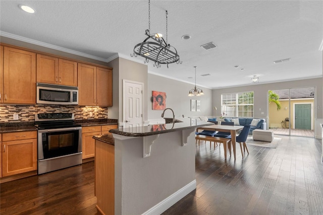 kitchen with dark wood-type flooring, a center island with sink, hanging light fixtures, appliances with stainless steel finishes, and a notable chandelier