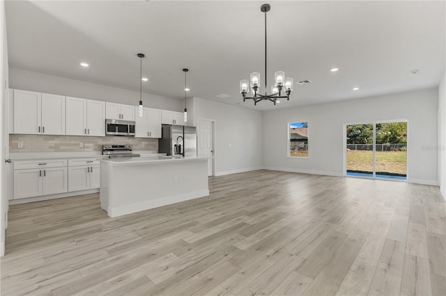 kitchen with hanging light fixtures, appliances with stainless steel finishes, a kitchen island with sink, white cabinets, and light wood-type flooring