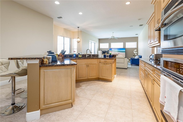 kitchen with appliances with stainless steel finishes, light brown cabinetry, dark stone counters, a kitchen island, and hanging light fixtures