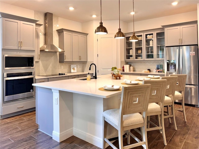 kitchen with sink, wall chimney exhaust hood, stainless steel appliances, dark hardwood / wood-style flooring, and an island with sink