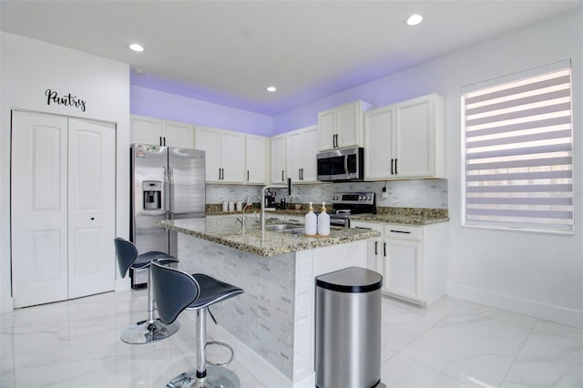 kitchen with light stone countertops, white cabinetry, stainless steel appliances, an island with sink, and a breakfast bar