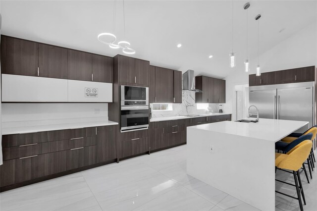 kitchen featuring wall chimney exhaust hood, built in appliances, pendant lighting, vaulted ceiling, and a kitchen island with sink