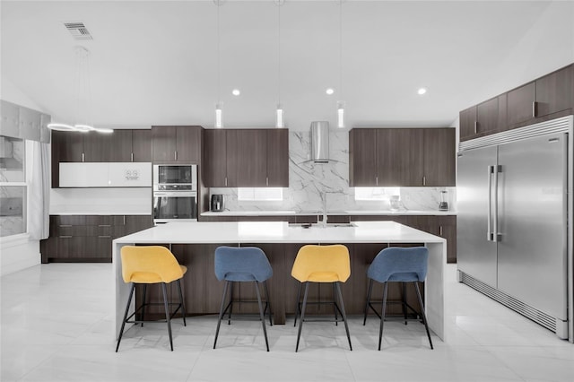 kitchen with built in appliances, a center island with sink, wall chimney range hood, and hanging light fixtures