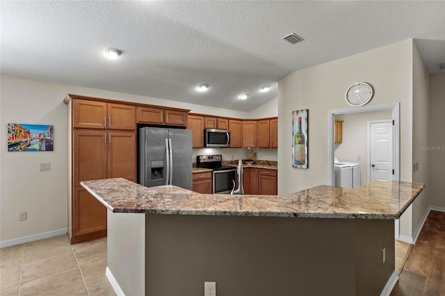 kitchen featuring appliances with stainless steel finishes, a kitchen island with sink, light stone counters, washer and dryer, and vaulted ceiling