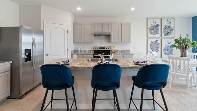 kitchen with gray cabinetry, sink, stainless steel appliances, light stone counters, and a center island with sink