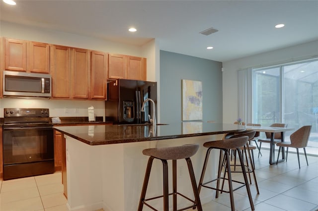 kitchen with a kitchen breakfast bar, dark stone counters, a kitchen island with sink, black appliances, and light tile patterned floors